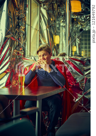 Man dressed as superhero pouring beer into glass, sitting in modern cafe with reflective walls, merging routine life with whimsical fantasy in vibrant surroundings. 121649792