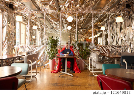 Superhero in bright cape sits in futuristic cafe showcasing pause in heroic duties to embrace leisure, connecting fantasy with human simplicity. Superhero in bright cape sits in futuristic cafe showcasing pause in heroic duties to embrace leisure, connecting fantasy with human simplicity. 121649812