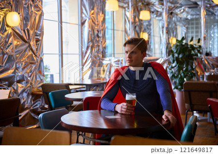 Superhero man sits with a glass of beer in reflective, metallic cafe, contemplating life's challenges, merging fantasy with moment of human relaxation. 121649817