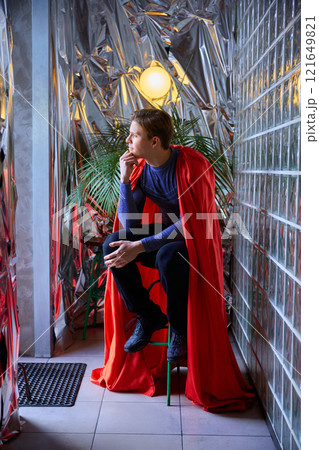 Superhero-dressed man sits by metallic wall, thoughtful gaze reflecting inner strength, futuristic vibes merging past inspiration and modern moments. 121649821