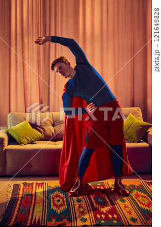 Superhero costume-wearing man bends sideways, performing a focused stretch, blending active routine with inner strength at home. 121649828