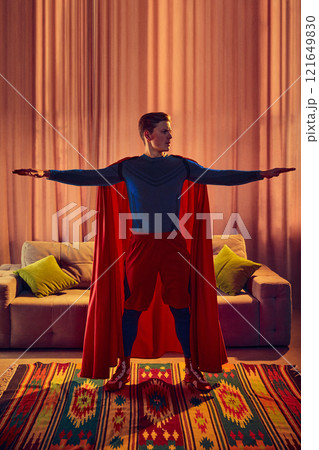 Man dressed as superhero stretches arms wide, training at home, to keep your body and spirit healthy, against warm tones of cozy living room. 121649830