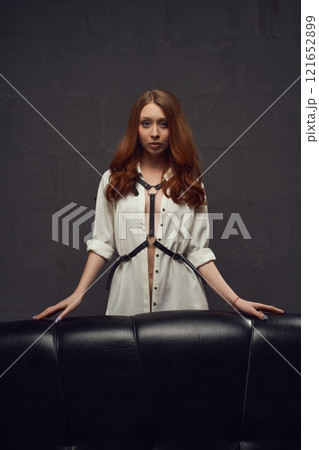 Slim woman in leather harness with black wings standing behind leather sofa Slim woman in leather harness with black wings standing behind leather sofa 121652899