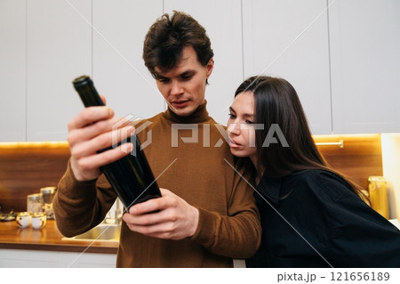 Couple examines a wine bottle in a modern kitchen during evening Couple examines a wine bottle in a modern kitchen during evening 121656189