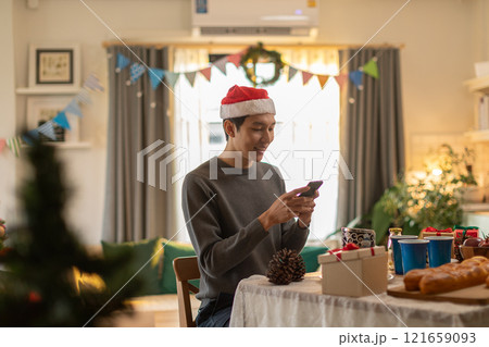 Man in Santa hat and casual sweater smiling while using smartphone at festively decorated table. Room is adorned with Christmas decorations, including garlands and colorful banners, as holiday treats 121659093