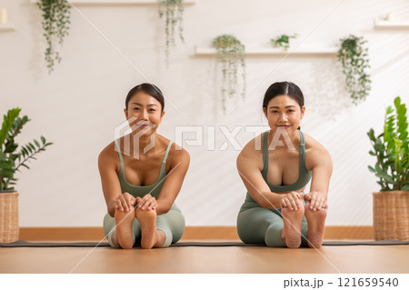 Two Asian women perform seated yoga stretch, bending gracefully to touch their feet showcasing flexibility and focus. They are dressed in matching green activewear with backdrop of natural plants 121659540