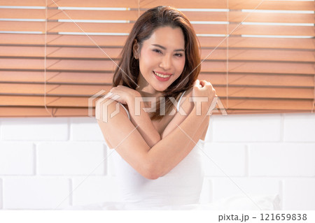 Cheerful Asian woman in white tank top, embracing herself with a warm and friendly smile. The background features wooden blinds, adding a cozy Cheerful Asian woman in white tank top, embracing herself with a warm and friendly smile. The background features wooden blinds, adding a cozy 121659838