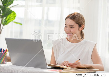 Smiling woman working on laptop at home, looking focused and productive during the day with natural light streaming in through windows. 121661265
