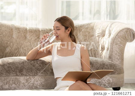 Woman sitting on the floor, drinking water while reading a book in a cozy and bright living room with a sofa in the background. 121661271