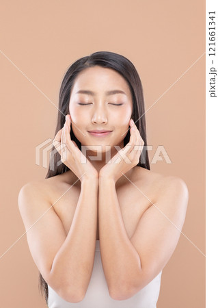 Serene and beautiful Asian young woman with flawless skin and long black hair. Gently touching her face with both hands, eyes closed, and smiling softly on soft peach color background 121661341