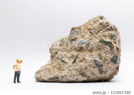 Miniature people construction worker stands next to a massive rock, scratching his head in contemplation 121661380