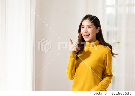 Smiling young woman in yellow sweater pointing upwards, expressing idea or inspiration, standing in brightly lit room. Smiling young woman in yellow sweater pointing upwards, expressing idea or inspiration, standing in brightly lit room. 121661539