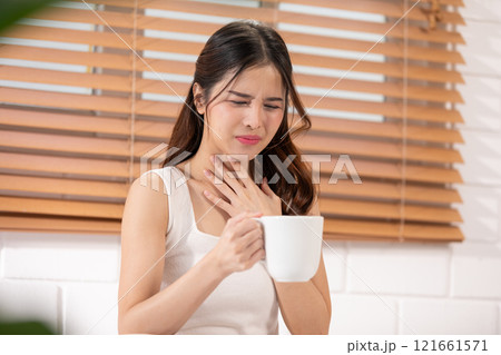 Asian woman in a white tank top holding a cup, showing discomfort and touching her throat, Health care concept Asian woman in a white tank top holding a cup, showing discomfort and touching her throat, Health care concept 121661571