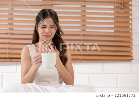 Asian woman in a white tank top holding a cup, showing discomfort and touching her throat, Health care concept Asian woman in a white tank top holding a cup, showing discomfort and touching her throat, Health care concept 121661572