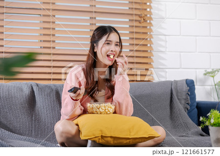 Asian young woman in pink cardigan and white top is comfortably seated on a couch, holding TV remote control in one hand and bowl of popcorn, Focused on the screen enjoying relaxed moment at home Asian young woman in pink cardigan and white top is comfortably seated on a couch, holding TV remote control in one hand and bowl of popcorn, Focused on the screen enjoying relaxed moment at home 121661577