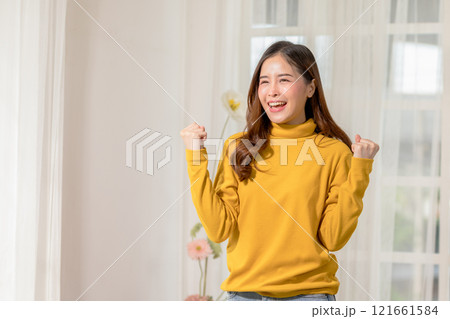Happy woman in yellow sweater celebrating success indoors, showing joy and excitement with raised fists. Bright interior background. Happy woman in yellow sweater celebrating success indoors, showing joy and excitement with raised fists. Bright interior background. 121661584