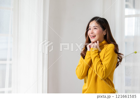 Joyful woman in yellow sweater expressing happiness and surprise indoors near white curtains in bright, minimalist room. Joyful woman in yellow sweater expressing happiness and surprise indoors near white curtains in bright, minimalist room. 121661585