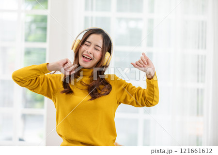 joyful Asian woman wearing a yellow sweater and headphones, dancing and enjoying music in an indoor setting. Her eyes are closed, and she is smiling widely 121661612