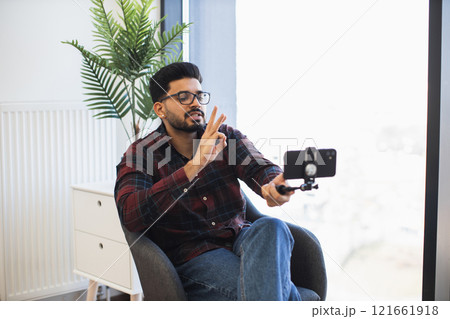 Young Indian man sitting indoors recording video content with smartphone. Wearing casual clothing, making gestures. Bright setting, natural light highlighting creative process and focused expression. Young Indian man sitting indoors recording video content with smartphone. Wearing casual clothing, making gestures. Bright setting, natural light highlighting creative process and focused expression. 121661918