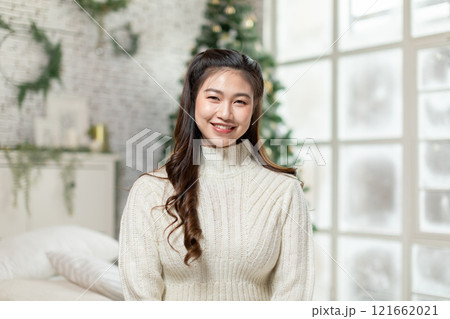 Cheerful Asian young woman smiles warmly, dressed in cozy sweater, standing in beautifully decorated holiday room. Festive setting includes Christmas tree and soft winter-themed decorations 121662021
