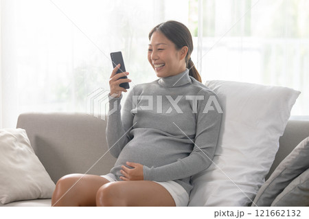 Asian pregnant woman in a grey turtleneck sweater is seated on a comfortable couch, smiling as she looks at her smartphone Asian pregnant woman in a grey turtleneck sweater is seated on a comfortable couch, smiling as she looks at her smartphone 121662132