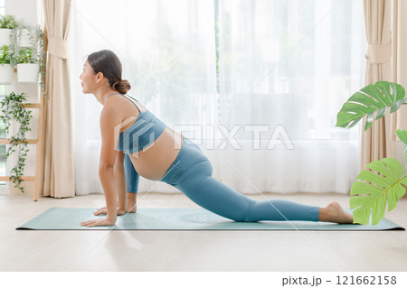 Pregnant woman practicing yoga at home, demonstrating a low lunge pose on a mat in a bright, airy room with indoor plants. 121662158