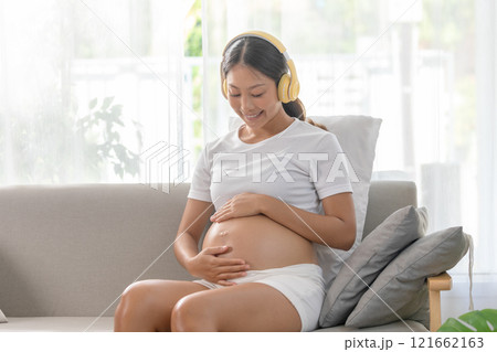 Pregnant woman sitting on couch, listening to music with headphones, hand on belly, wearing comfortable clothes, relaxed and happy at home. 121662163