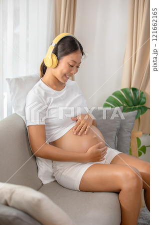 Pregnant woman wearing headphones sitting on couch, smiling and holding her belly, enjoying music in a cozy living room setting. 121662165