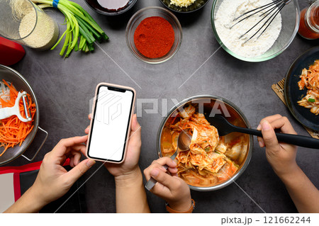 Overhead view of women preparing Kimchi with smartphone for recipe guidance on a dark countertop surrounded by fresh ingredients Overhead view of women preparing Kimchi with smartphone for recipe guidance on a dark countertop surrounded by fresh ingredients 121662244