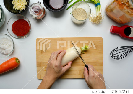 Above view of hands slicing daikon radish on cutting board surrounded by ingredients for making kimchi Above view of hands slicing daikon radish on cutting board surrounded by ingredients for making kimchi 121662251