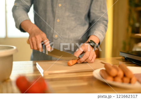 Cropped shot of man preparing simple meal in a kitchen, cutting sausages on a wooden cutting board. 121662317