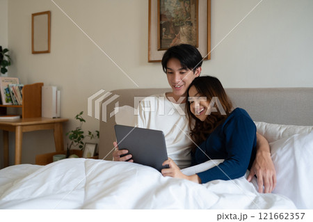 Loving adult couple using digital tablet under warm blanket in a cozy bedroom 121662357