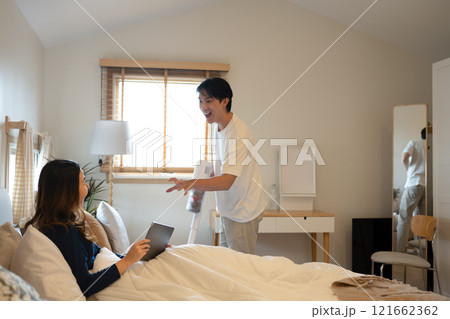 Happy young woman using digital tablet on bed while her husband vacuuming floor beside Happy young woman using digital tablet on bed while her husband vacuuming floor beside 121662362