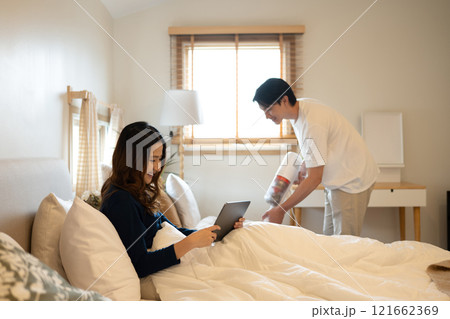 Cheerful woman using digital tablet on bed while her husband vacuuming floor beside 121662369