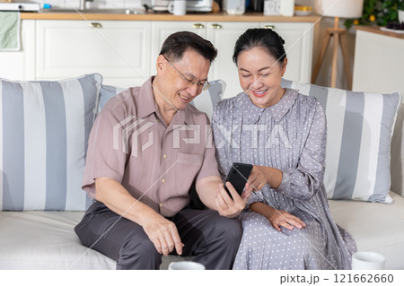 Asian senior couple sitting on couch, happily looking at smartphone screen. They are smiling and sharing moment of connection, showcasing the joy of exploring technology together in cozy home setting 121662660
