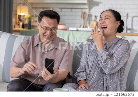 Joyful Asian senior couple sitting on couch, sharing hearty laugh while looking at smartphone. Highlighting their enjoyment of technology and the warmth of their relationship in cozy home environment 121662672