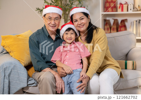 Joyful Asian family of three, including grandparents and child wearing Santa hat, sits together on couch, smiling warmly. Festive scene set indoors with Christmas decorations in background Joyful Asian family of three, including grandparents and child wearing Santa hat, sits together on couch, smiling warmly. Festive scene set indoors with Christmas decorations in background 121662702