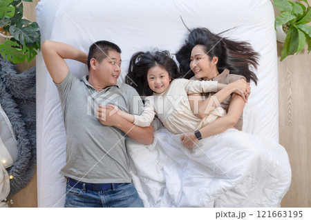 Asian joyful family scene with father, mother, and daughter lying together on bed. They are smiling and looking at each other, sharing moment of closeness and warmth 121663195