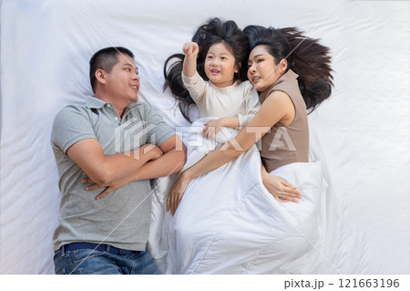 Asian joyful family scene featuring afather, mother, and their young daughter lying comfortably on a bed. The family is enjoying a peaceful and intimate moment, with the child pointing excitedly Asian joyful family scene featuring afather, mother, and their young daughter lying comfortably on a bed. The family is enjoying a peaceful and intimate moment, with the child pointing excitedly 121663196
