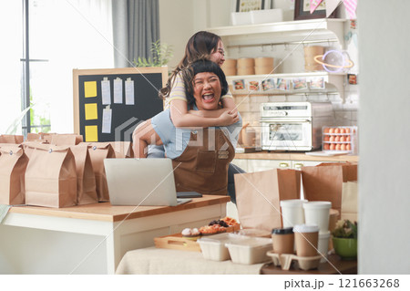 Two cheerful chefs share playful moment in cozy kitchen filled with food items and takeaway bags. One chef gives the other piggyback ride, both laughing heartily. The kitchen is warm and inviting 121663268