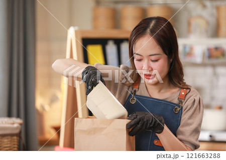 Asian young female chef, wearing apron and gloves, carefully packs food into paper bag in kitchen. Chef focused expression highlights her attention detail and care in preparing the meal for takeaway 121663288