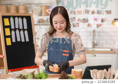Asian young female chef, wearing blue apron and gloves, slices bread in vibrant kitchen environment. Fresh ingredients visible on counter, emphasizing healthy and creative culinary approach 121663289