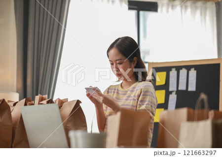 Focused Asian woman carefully managing food delivery orders in well-organized workspace. Surrounded by paper bags and working with notes. business management concept Focused Asian woman carefully managing food delivery orders in well-organized workspace. Surrounded by paper bags and working with notes. business management concept 121663297