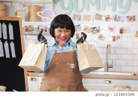 Cheerful Asian chef in brown apron and black gloves proudly holds up two takeaway bags, ready for delivery. The kitchen background is vibrant and cozy. Small business concept 121663306