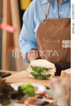 close-up view of a freshly prepared sandwich placed on a wooden cutting board. The sandwich, filled with lettuce, ham, and cheese, is cut in half, showcasing the layers of fresh ingredients 121663307