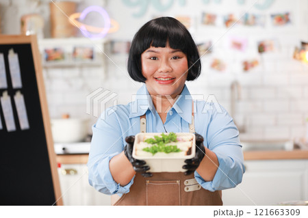 Cheerful Asian chef wearing a brown apron and black gloves presents a freshly prepared sandwich box, smiling warmly at the camera. Small Business Concept Cheerful Asian chef wearing a brown apron and black gloves presents a freshly prepared sandwich box, smiling warmly at the camera. Small Business Concept 121663309