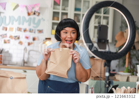 Enthusiastic Asian Content Creator Presenting Food Item in Modern Kitchen Setup, including ring light and smartphone, to film culinary tutorial or review. Small Business Concept 121663312