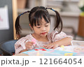 Asian young girl with Down syndrome wearing pigtails, is immersed in arts and crafts project at table. She holds piece of colored paper with focus and concentration, surrounded by crafting materials 121664054