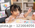 Asian young girl with Down syndrome wearing pigtails, is immersed in arts and crafts project at table. She holds piece of colored paper with focus and concentration, surrounded by crafting materials 121664056