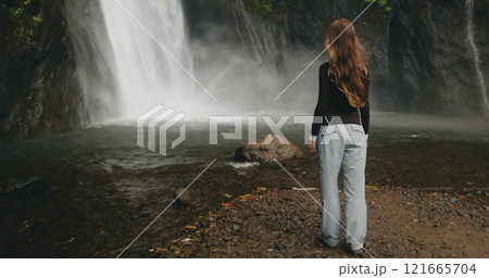 Female hiker is admiring a powerful waterfall cascading down a rocky cliff in a lush tropical rainforest. She is enjoying the peace and serenity of nature during her adventure travel in indonesia 121665704